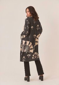 Mujer vestida con un abrigo largo negro con patrones florales beiges, pantalones negros y zapatos de tacón negro, de pie con las manos en los bolsillos.