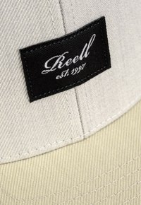Beige Kappe aus strukturiertem Stoff mit flacher Krempe. Schwarzes Etikett mit weißem, geschwungenem Text "Reell est. 1997", der auf das Vorderpanel gestickt ist.