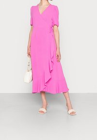 Vestido envolvente fucsia con mangas abullonadas, dobladillo con volantes y cinturón de lazo. Combinado con sandalias de tacón blancas y un bolso blanco. Textura de tela suave.