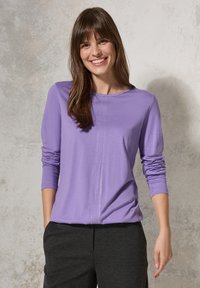 Lila Langarmshirt aus weichem Stoff, mit Rundhalsausschnitt und einer Mittelnähtedetail, kombiniert mit dunkelgrauen Hosen.