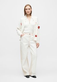 Hvit strikket cardigan med røde blomsterapplikasjoner, kombinert med off-white bukser med vide ben og kontrastfargede sømmer. Svarte sko fullfører antrekket.