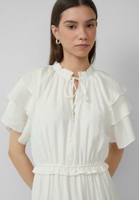 Witte, lichte jurk met gerimpelde mouwen, getailleerde taille en een ronde halslijn met een strikdetail en een subtiele knoopsluiting.