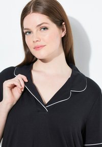 Zwart top met witte biezen, met een kleine kraag en een V-hals ontwerp. De stof lijkt zacht en lichtgewicht.