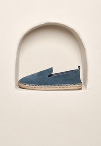 Blauer Wildleder-Slipper mit gewebter Jutesohle und sichtbaren Nähten. Präsentiert in einer versenkten Nische vor einem glatten weißen Hintergrund.