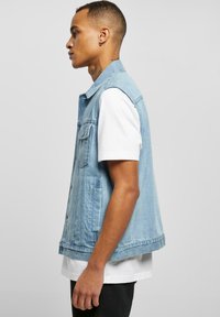 Jachetă din denim fără mâneci, de culoare albastru deschis, având guler clasic, buzunare frontale și un design drept. Purtați-o peste un tricou alb.
