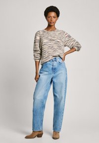 Maglione lavorato a maglia con strisce multicolori; vestibilità ampia. Jeans chiari a vita alta; gamba larga. Stivali alla caviglia marroni. Sfondo neutro.