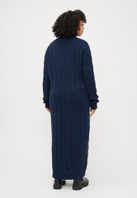 Eine Person mit lockigem Haar trägt ein langes, navyblaues Strickkleid mit einem Kabelmuster, von hinten gesehen, vor einem schlichten weißen Hintergrund.