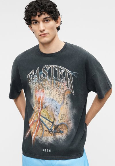 Jeune homme aux cheveux bouclés foncés portant un T-shirt graphique noir avec un design fissuré, un éclair et un vélo dans des couleurs atténuées.