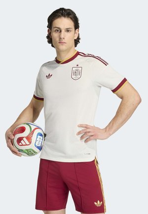 Mann in weiß-rotem Spanien-Fußballtrikot und roten Shorts hält mit einer Hand an der Hüfte einen mehrfarbigen FIFA-Fußball.
