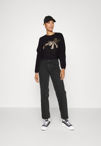 Pull noir avec motif de nœud doré, associé à un jean noir taille haute et des baskets noir et blanc. Le mannequin porte une casquette noire.