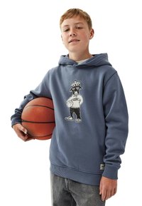 Sininen puuvillahuppari, jossa on graafinen printti aurinkolaseja ja kruunua kantavasta koirasta, yhdistettynä harmaisiin farkkuihin ja koriin.