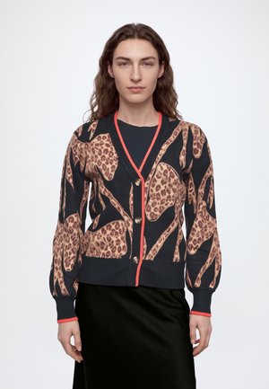 Modèle portant un cardigan à imprimé léopard sur un haut foncé et une longue jupe noire, avec des escarpins noirs et un sac à main, devant un fond de studio blanc.