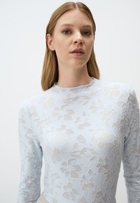 Top lungo blu chiaro a maniche lunghe realizzato in pizzo con motivi floreali. Presenta un collo alto e una silhouette aderente, mettendo in mostra texture stratificate.