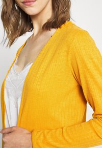 Žlutý žebrovaný cardigan s texturovaným povrchem, s otevřeným předním designem a dlouhými rukávy, vrstvený přes bílé tričko s krajkovým lemem.