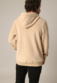 Sweat à capuche en polaire beige avec un cordon de serrage, des manches longues et une poche avant. Tissu texturé, coupe décontractée et détail en bord-côtes.
