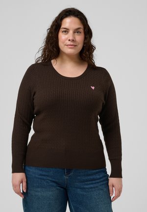 Femme aux cheveux bruns bouclés portant un pull torsadé brun foncé avec un petit cœur rose et un jean bleu, debout devant un fond uni.