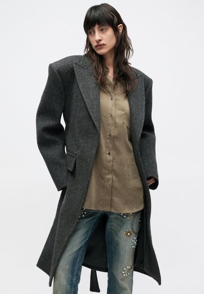 Kvinde iført stor, mørkegrå frakke, olivengrøn skjorte med knapper og blå jeans med dekorative detaljer, kigger sidelæns mod en ensfarvet baggrund.