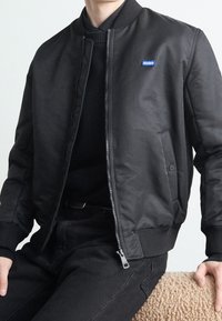 Schwarze Bomberjacke aus glattem Stoff, mit einem Frontreißverschluss, gerippten Bündchen und einem blauen "HUGO"-Logo auf der linken Brust.