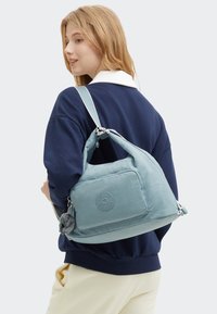 Soft blue nylon bag with a rounded shape, front pocket, and a textured logo. Features a detachable pom-pom keychain and adjustable strap.