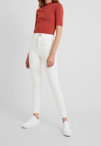 Kvinna klädd i en roströd kortärmad topp, högmidjade vita skinny jeans och vita sneakers, stående mot en enkel bakgrund.