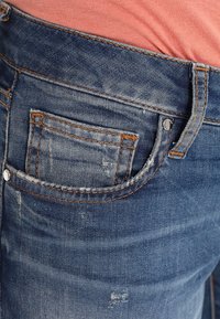 Vaqueros de mezclilla en azul oscuro con un ligero desgastado y efecto de arrugas. Presentan un bolsillo trasero con costura naranja y un detalle de remache plateado.