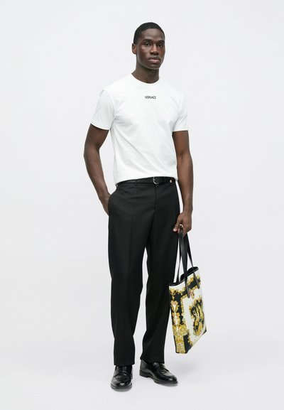 Biała koszulka z krótkim rękawem z logo „VERSACE”, czarne dopasowane spodnie, błyszczące czarne buty oraz torebka typu tote z wzorem w kolorach czarnym, białym i złotym.