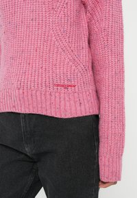 Rosa strikkegenser med strukturelt mønster, ribbede mansjetter og subtile flerfargede prikker. Merkevaren er brodert i rødt.