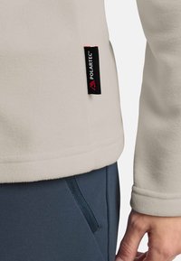 Giacca in fleece beige chiaro con etichetta Polartec nera e rossa, abbinata a pantaloni blu scuro con tasca laterale con zip.