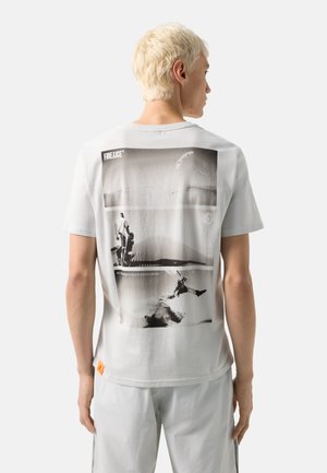 Person med kort blondt hår bærer en hvid t-shirt med sort-hvide ørken- og paraglidingfotoer på ryggen.