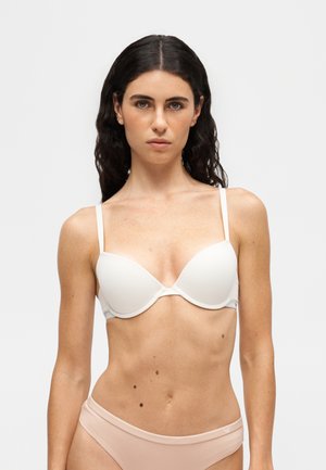Donna con capelli lunghi e scuri che indossa un reggiseno bianco e biancheria intima beige chiaro, in piedi davanti a uno sfondo chiaro e uniforme.