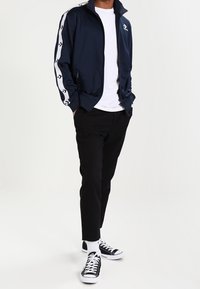 Chándal de navy con franjas blancas en los laterales, cierre de cremallera y cuello alto; combinado con pantalones negros y zapatillas clásicas negras. Camiseta blanca debajo.