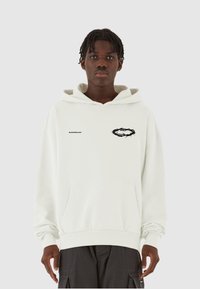 Weißer Kapuzenpullover mit einem schwarzen Grafiklogo und Text auf der linken Brust, Kängurutasche und gerippten Bündchen. Modell trägt dunkle Cargo-Hosen.