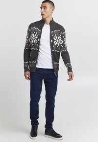 Grauer gestrickter cardigan mit weißen Schneeflocken-Mustern, Reißverschluss und gerippten Bündchen, kombiniert mit einem weißen T-Shirt und dunklen Jeans.