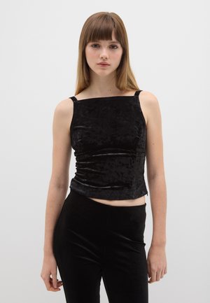 Crop top en velours noir avec un décolleté carré et des fines bretelles. Le tissu a une texture lisse et un léger éclat. Associé à un pantalon noir.