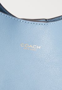 Svijetloplava koža s glatkom teksturom, s zlatno urezanim logotipom "COACH NEW YORK" i kontrastnim šavovima.