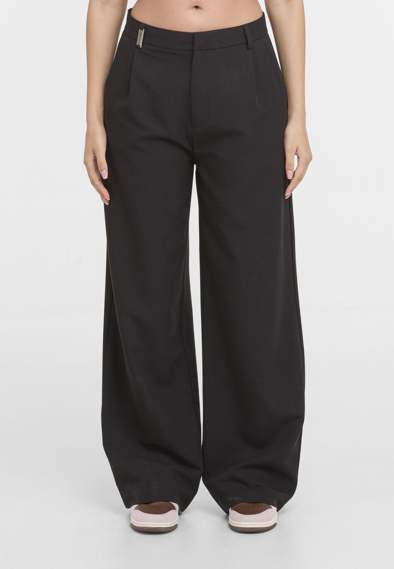 Pantalon noir à jambes larges avec devant plat, poches latérales et passants de ceinture, porté avec des baskets roses et brunes sur un fond blanc.