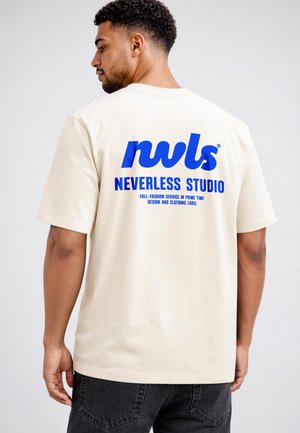 Mann trägt cremefarbenes T-Shirt mit blauem "nwls NEVERLESS STUDIO"-Text auf dem Rücken, kombiniert mit schwarzen Jeans, steht vor einfachem hellem Hintergrund.
