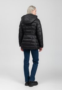 Zwarte gewatteerde jas met capuchon, met horizontale stiknaden, ritssluiting en een strakke textuur. Draagt bij donkere spijkerbroek en stevige zwarte laarzen.