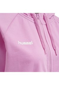 Rosa zip-up hoodie i mjukt tyg med en texturerad yta. Har en vit logotyp på vänster bröst och dragsnoddar för justering på huvan.