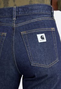 Blugi din denim de culoare albastră închisă au un croi drept, cusături vizibile de culoare galbenă și o etichetă albă a brandului pe buzunarul din spate.