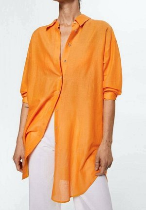 Personne portant une chemise orange longue et ample boutonnée avec les manches retroussées et un pantalon blanc sur un fond uni.
