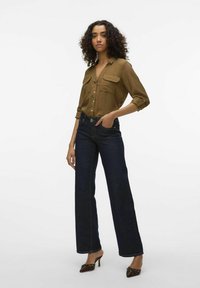 Camisa marrón de botones con dos bolsillos en el pecho, combinada con jeans de pierna ancha de color azul oscuro. El modelo lleva unos zuecos marrones de tacón con Acabados decorativos.