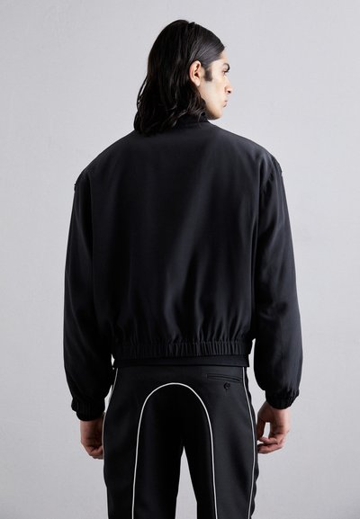 Jachetă bomber neagră cu talie și manșete elastice, având o textură netedă și un design minimalist, fără modele sau logo-uri vizibile.