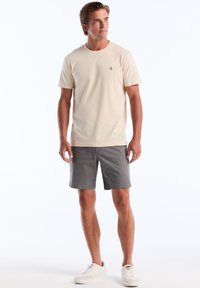 T-shirt en coton beige avec un petit logo bleu marine, associé à un short gris et des baskets blanches. Design simple et coupe décontractée.