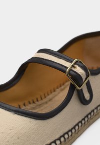 Espadrille en toile beige avec bordure noire, bride noire et beige ornée d'une boucle dorée, et coutures noires détaillées le long de la semelle.