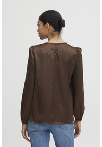 Blusa di satin marrone con maniche a sbuffo lunghe, chiusura con bottoni sul retro e una vestibilità comoda. Abbinata a jeans in denim blu.