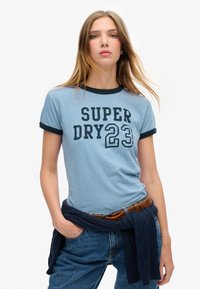 Lichtblauwe t-shirt met korte mouwen, marineblauwe geribde kraag en mouwen, met "SUPER DRY 23" in vetgedrukte zwarte letters. Casual pasvorm, gedragen met denim.