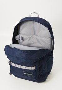 Granatowy plecak z zamykanym main compartment, szare wnętrze, przednia kieszeń z pasekowym akcentem oraz uchwyt do noszenia u góry.