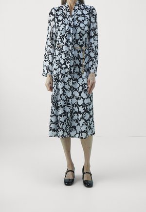 Femme portant une robe midi fleurie noire et blanche avec une ceinture chaîne dorée et des chaussures Mary Jane noires, debout sur un fond blanc uni.