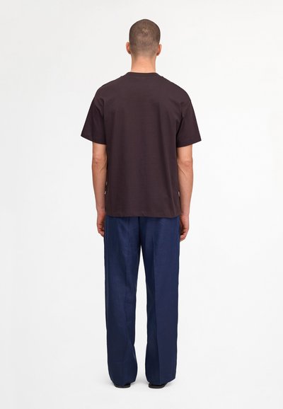 Homme debout, tourné vers l'arrière, portant un t-shirt noir à manches courtes et un pantalon ample bleu marine sur fond blanc uni.